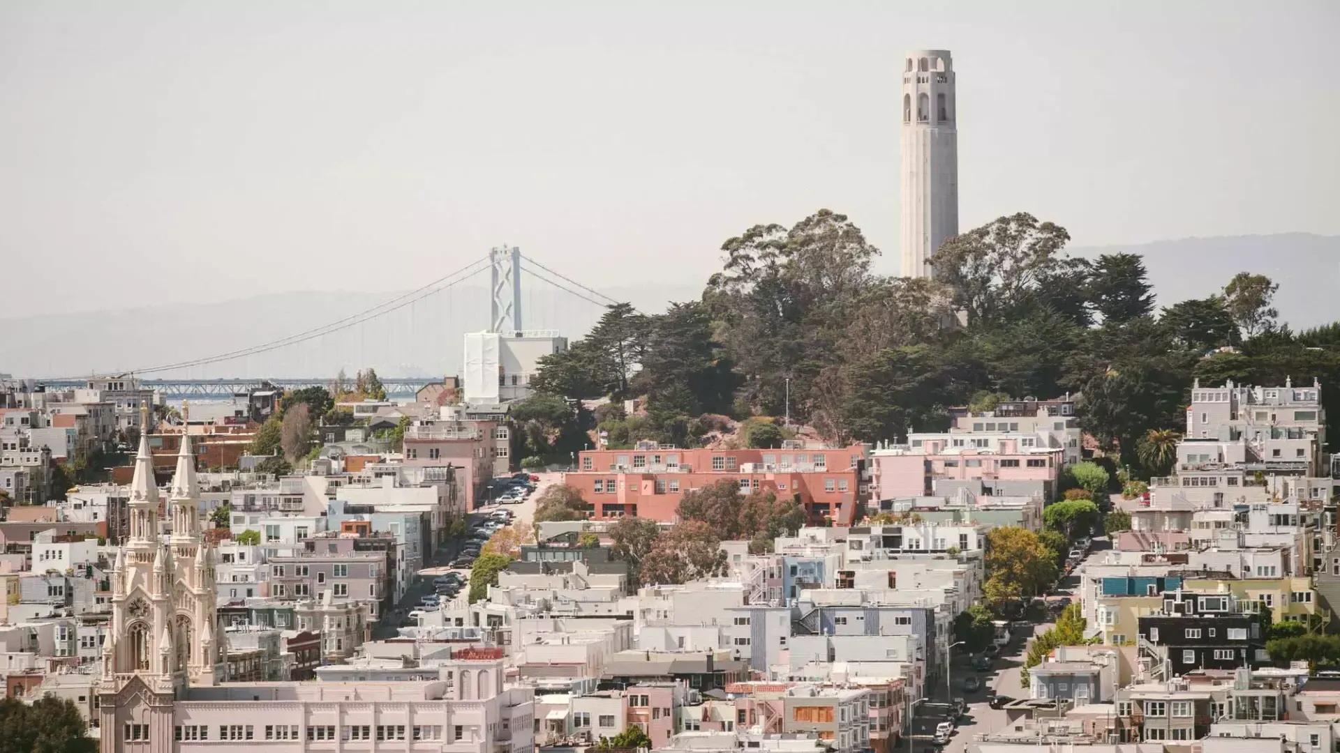 Abgebildet ist der Coit Tower in San Francisco mit der Bay Bridge im Hintergrund und einem mit Häusern bedeckten Hügel im Vordergrund.